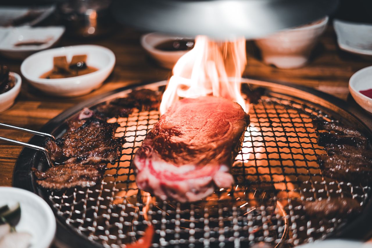 services-01 Close-up of a steak grilling with flames on a barbecue, surrounded by dishes.