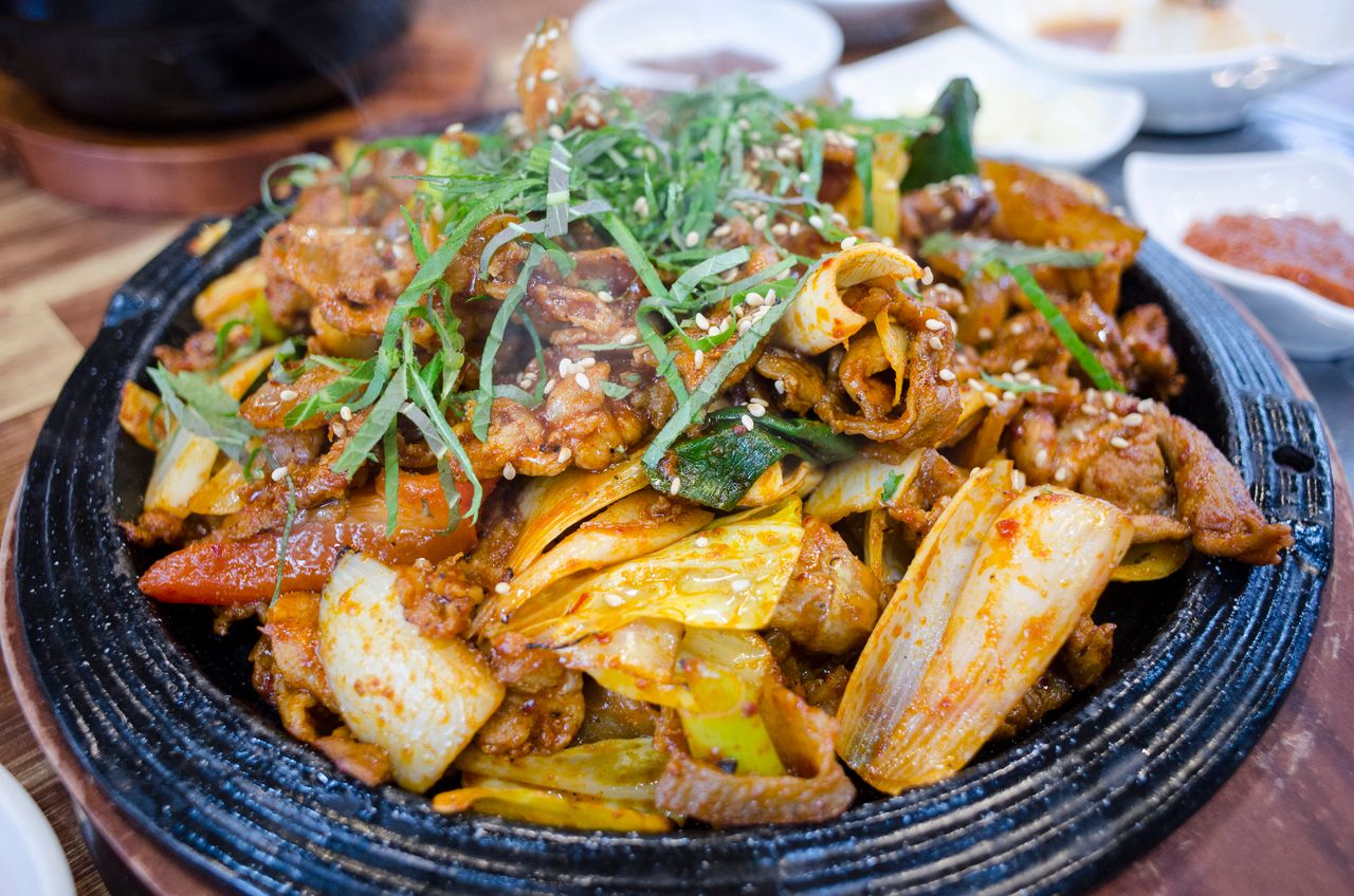 heros-img Close-up of a delicious Korean BBQ pork dish with vegetables on a sizzling plate.